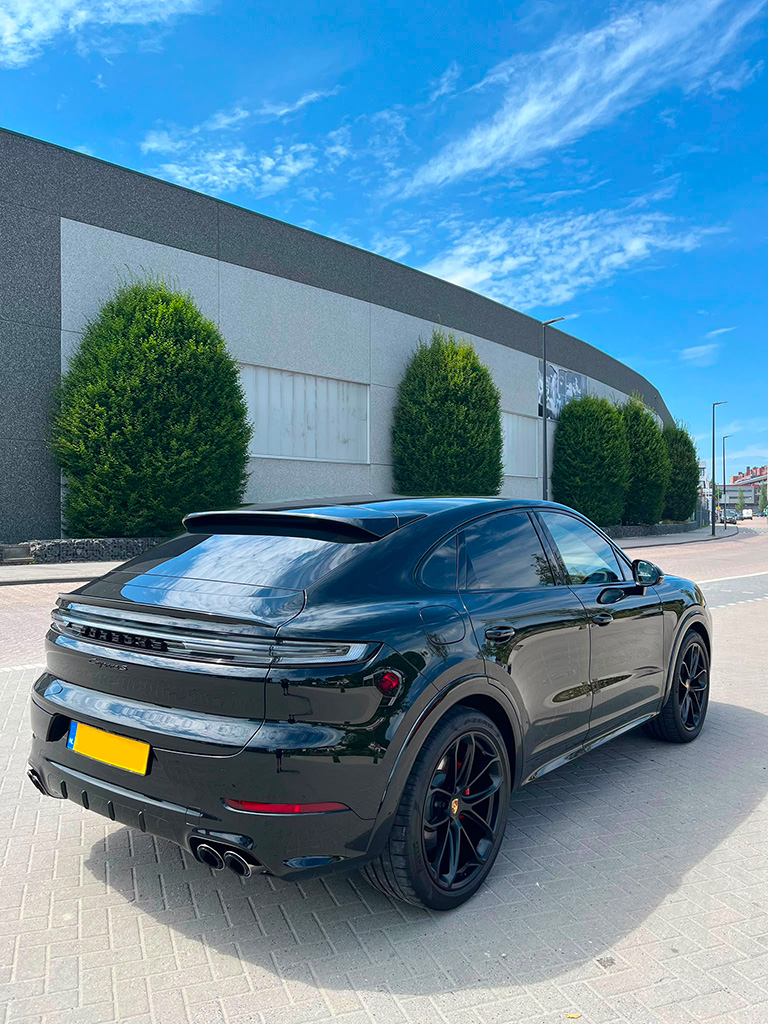 Schuin achteraanzicht van een glanzend zwarte Porsche Cayenne S Coupé geparkeerd op een klinkerstraat onder een blauwe lucht